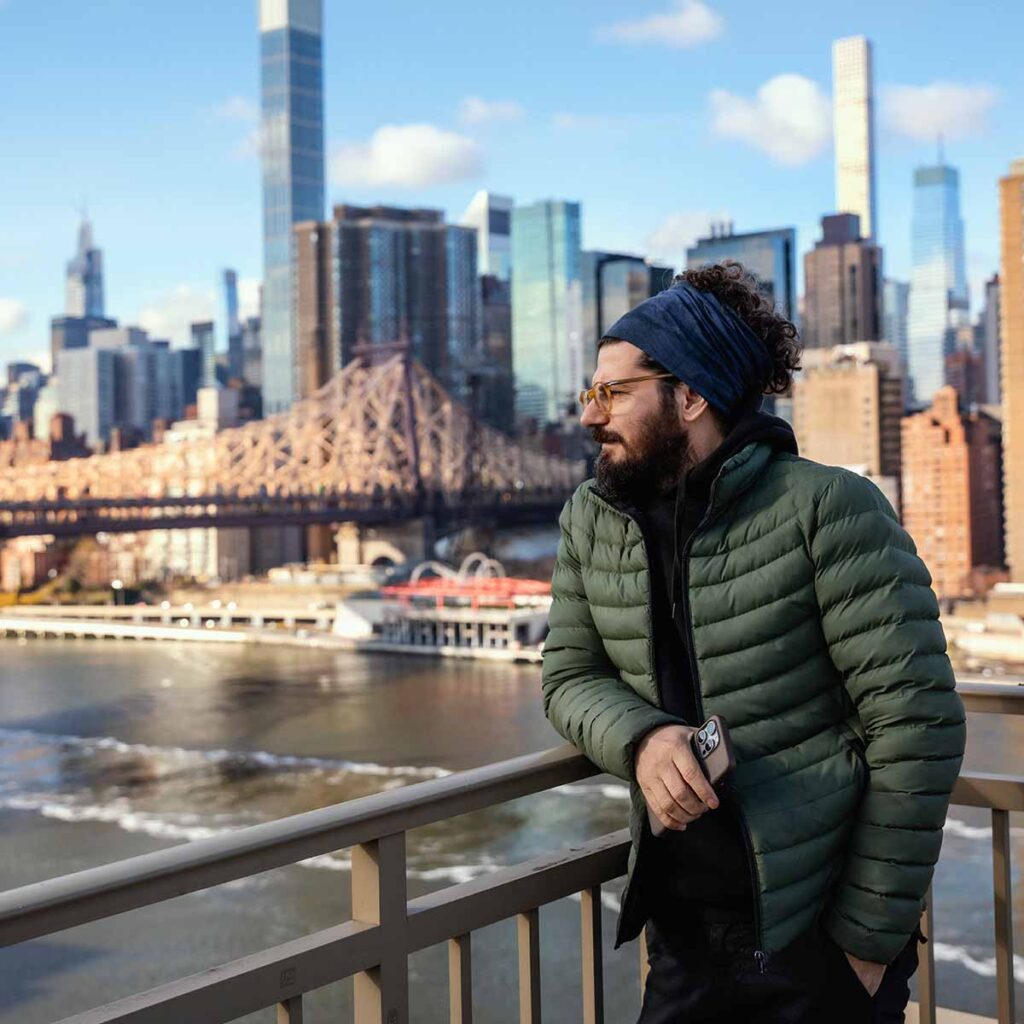 Man in green winter jacket on bridge with NYC in background